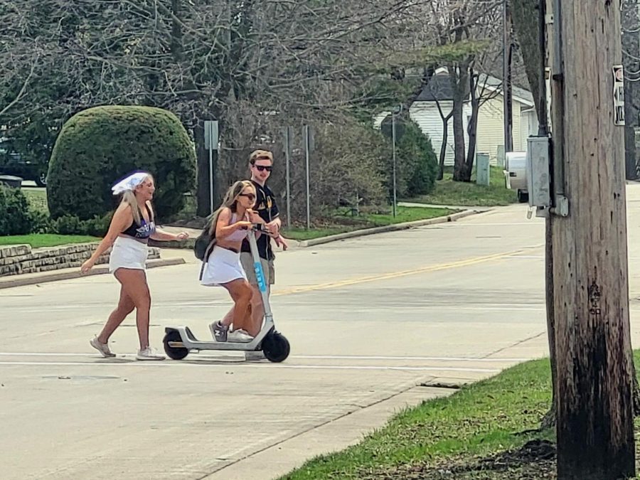 Three friends make their way across the street with one on an electric scooter.
