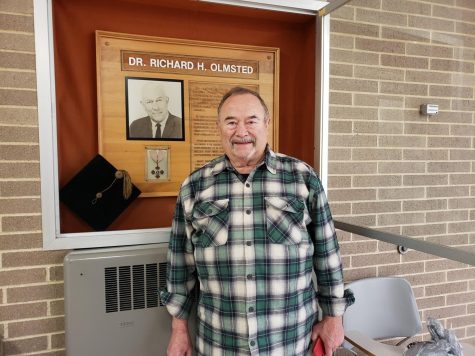 Jim Olmsted son of Dr. Richard Olmsted poses in front of a plaque in his fathers honor outside lecture hall 100 of Heide Hall.