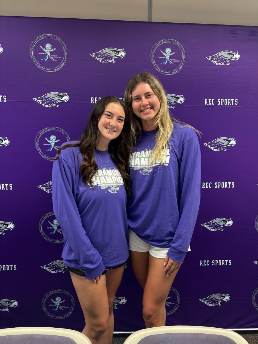 Brynn Fuerstenberg (right) poses with Cailyn Kramer (left) as they smile at the camera, wearing an intramural championship shirt in front of a photo wall that is decorated with the University Warhawk and rec sports logo. (Submitted by Brynn Fuerstenberg)
